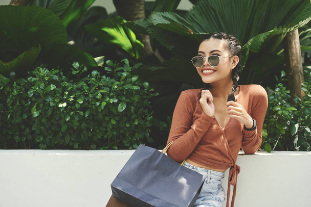 Mujer elegante sonriente en gafas de sol de pie con bolsas de compras y disfrutar del buen tiempo con árboles tropicales en el fondo
 - Foto, imagen