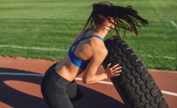 Rear view image of athlete fit young woman doing tire-flip outdoors on the stadium. Copy space for advertising. Sportswoman doing workout exercises outside. People, lifestyle and sport - Foto, Imagen