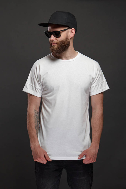 Hipster handsome male model with beard wearing white blank t-shirt and a baseball cap with space for your logo or design in casual urban style - Photo, Image