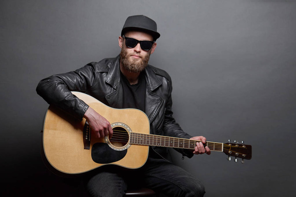 Guitar player singing in music studio. Hipster guitar player with beard and black clothes playing the acoustic guitar - Foto, Imagen