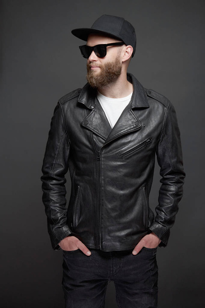Man wearing leather biker jacket or asymmetric zip jacket with black cap, jeans and sunglasses. Handsome hipster man over gray background - Foto, Imagen
