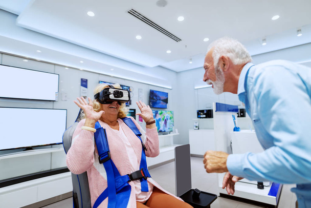 Симпатичні посміхаючись кавказька старша пара випробувати технологію віртуальної реальності. Жінка сидить у кріслі і носить VR окуляри. Інтер'єр магазину Tech. - Фото, зображення