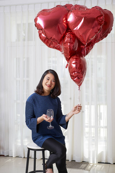 Portrait of young Asian woman sitting on chair holding red heart shaped balloons and empty wineglass and looking at camera - Foto, Bild