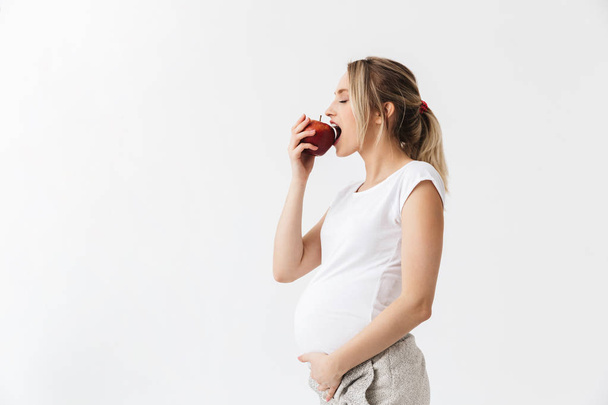 Beautiful young pregnant woman posing isolated over white wall background holding apple eat healthy food. - Фото, зображення
