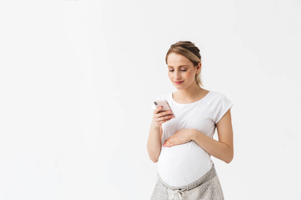 Happy young pregnant woman posing isolated over white wall background using mobile phone. - Zdjęcie, obraz