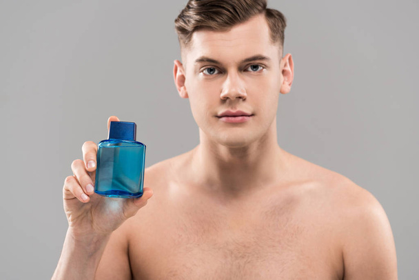 vista frontal del guapo joven desnudo sosteniendo botella de loción aislada en gris
 - Foto, imagen