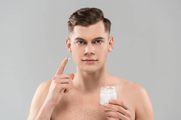 Vorderseite des nackten jungen Mannes mit Kosmetikbehälter und Anwendung Gesichtscreme isoliert auf grau - Foto, Bild