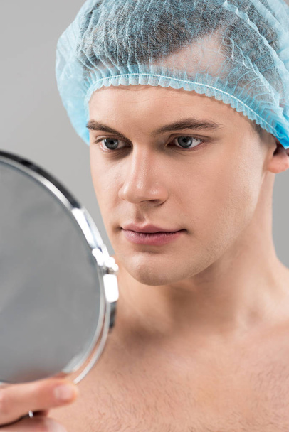 fiatal férfi orvosi sapkában nézi a szürke üvegen izolált tükröt. - Fotó, kép