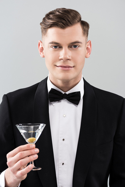 front view of smiling young man in formal wear with bow tie holding glass of cocktail isolated on grey - Foto, afbeelding