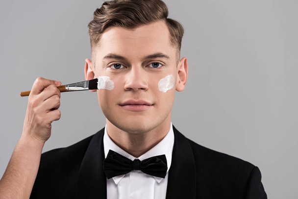 abgeschnittene Ansicht eines Kosmetikers, der Creme aufträgt und einen Mann in formeller Kleidung isoliert auf grau - Foto, Bild