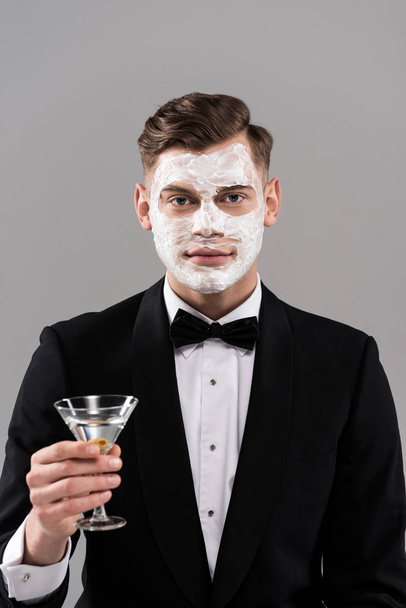 front view of man in formal wear with cream on face holding glass of cocktail isolated on grey - Fotoğraf, Görsel