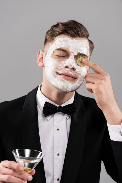 νεαρός άνδρας με επίσημη ένδυση με μάσκα προσώπου που κρατά ένα ποτήρι κοκτέιλ και απλώνει κομμένο αγγούρι στο μάτι που απομονώνεται στο γκρι - Φωτογραφία, εικόνα