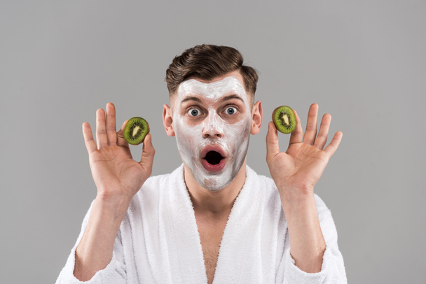 front view of shocked man in bathrobe holding cut kiwi isolated on grey - Foto, immagini