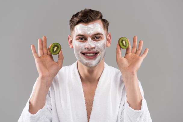 front view of smiling man in bathrobe holding cut kiwi isolated on grey - Photo, Image