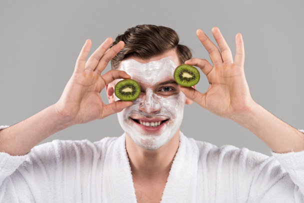 front view of smiling man in bathrobe holding cut kiwi isolated on grey - Фото, изображение