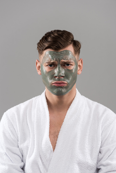 front view of sad young man in bathrobe with clay mask isolated on grey - Foto, immagini