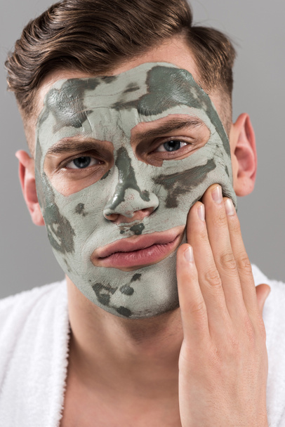 displeased young man with clay mask touching face isolated on grey - Foto, afbeelding
