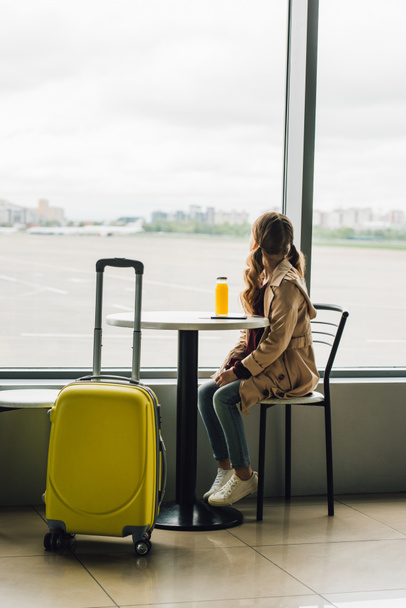 preteen kid sitting near yellow suitcase in waiting hall in airport and looking through window - Photo, image