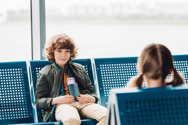 chlapec, který sedí na modrém sedadle, drží pas a dívá se na přítele v čekárně na letišti  - Fotografie, Obrázek