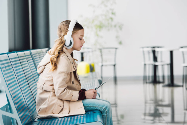 vista lateral da criança pré-adolescente sentado no assento azul, segurando passaporte e ouvindo música na sala de espera no aeroporto
  - Foto, Imagem