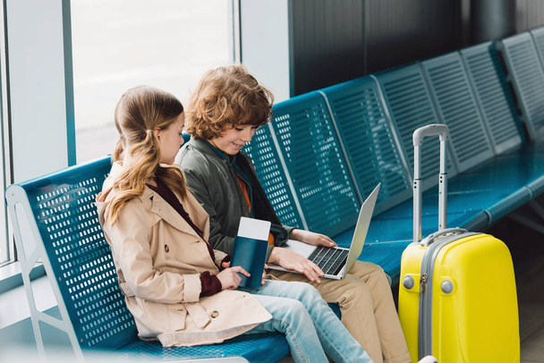 kinderen zittend op blauwe zitplaatsen in de buurt van gele koffer in wachtzaal in de luchthaven  - Foto, afbeelding