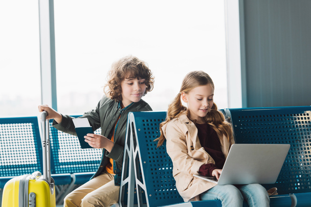Preteen Kid typen op laptop, terwijl Boy Holding paspoort en gele koffer in wachtzaal in de luchthaven - Foto, afbeelding