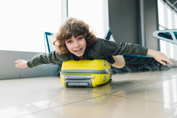 excited preteen kid lying on suitcase with outstretched hands in airport departure lounge - Foto, Imagem