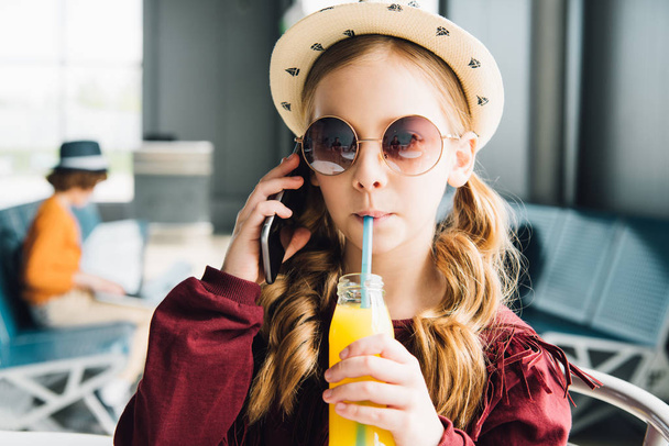 preteen kid drinking orange juice and talking on smartphone in waiting hall - Foto, Imagen