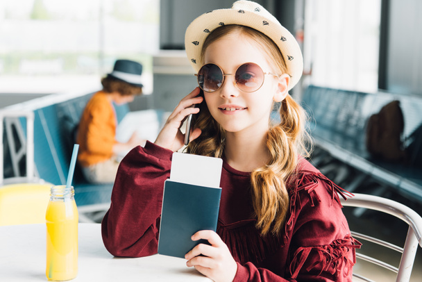 niño preadolescente sonriente sosteniendo pasaporte y hablando en el teléfono inteligente en la sala de espera
 - Foto, imagen