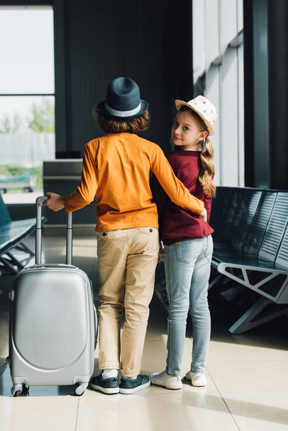 Вид сзади на мальчика-подростка с чемоданом, обнимающего ребенка-подростка в приемной
 - Фото, изображение