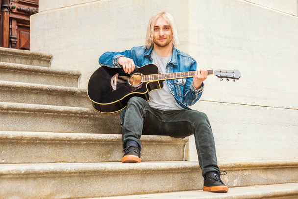 Молодой студент колледжа с длинными светлыми волосами, одет в синюю джинсовую куртку, серую рубашку, черные брюки, кроссовки, сидит на лестнице возле здания винтажного стиля в кампусе в Нью-Йорке, играет на гитаре
 - Фото, изображение