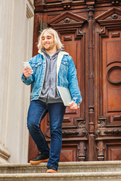 Jonge hipster student met lang blond haar, blauwe Denim jas, blauwe jeans, sneakers, laptop computer vasthouden, de trap af lopen vanuit New York, sms 'en, glimlachen - Foto, afbeelding