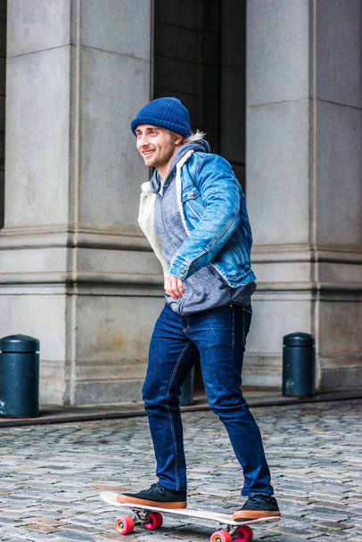 Patinaje en la calle en Nueva York. Joven estudiante universitario, vistiendo chaqueta azul de mezclilla, sombrero de punto, jeans, zapatillas, de pie en el monopatín en la calle vintage en el campus, mirando, patinando, sonriendo
 - Foto, Imagen