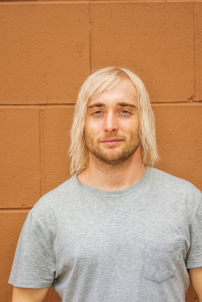 Portrait of Young Man in New York City. Young hipster college student with long blonde hair, wearing gray T shirt, standing against brown wall on street - Fotó, kép