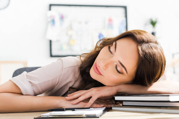 cheerful young woman with closed eyes lying near clipboard and pen on wooden desk - Photo, Image