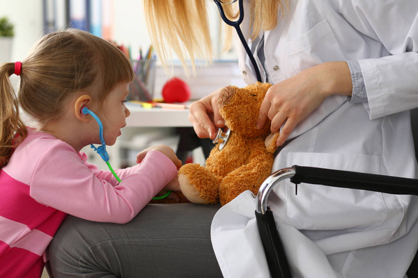Маленька дівчинка тримає в руках іграшковий стетоскоп, граючи з плюшевим ведмедем
 - Фото, зображення