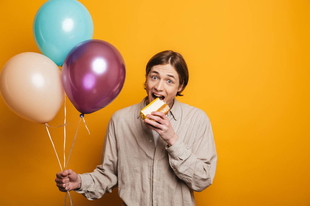 Happy handsome man in shirt holding balloons while eating cake - Foto, immagini
