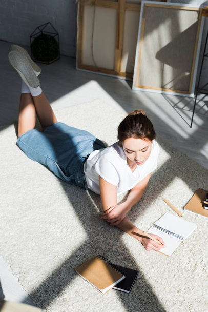 pretty young woman in white t-shirt lying on carpet and writing in notebook  - Zdjęcie, obraz