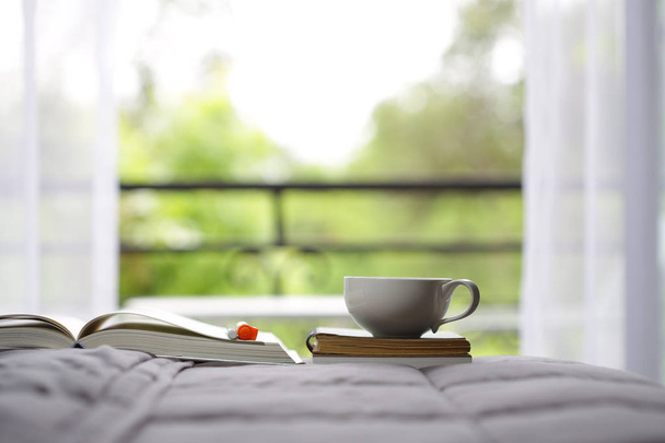 窓の景色を望むベッドの上のコーヒーカップとノート - 写真・画像