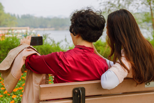 concepto de familia, tecnología y personas - feliz hija sonriente y madre mayor con teléfono inteligente sentado en el banco del parque y tomando selfie
. - Foto, Imagen