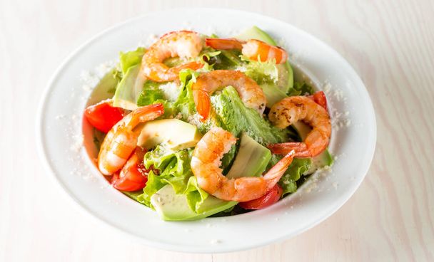Fresh shrimp salad made of tomato, ruccola, avocado, prawns, chicken breast, arugula, crackers and spices. Caesar salad in a white, transparent bowl on wooden background - Фото, зображення