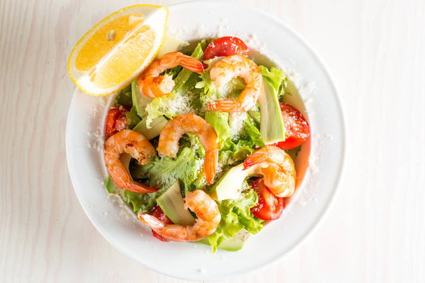 Fresh shrimp salad made of tomato, ruccola, avocado, prawns, chicken breast, arugula, crackers and spices. Caesar salad in a white, transparent bowl on wooden background - Fotografie, Obrázek