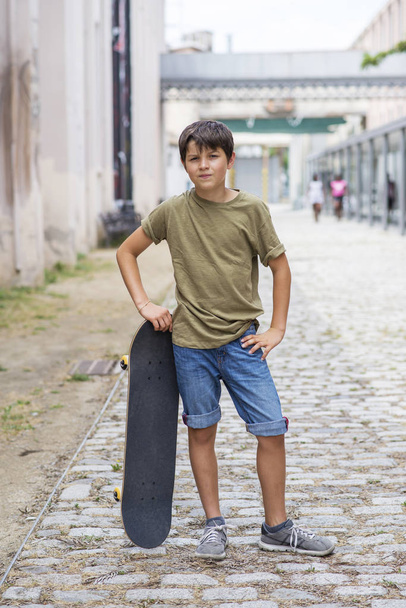 明るい日にカメラを見ている間にスケートボードを保持しながら、若い陽気な男の子が路上で立っている - 写真・画像