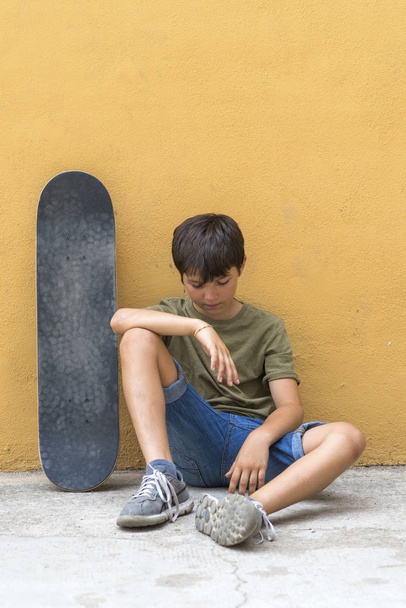 Vooraanzicht van een jonge jongen zittend op de grond leunend op een gele muur terwijl naar beneden kijken - Foto, afbeelding