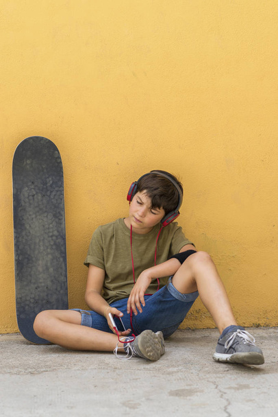 ヘッドフォンで音楽を聴きながら黄色い壁に座っている若いスケーターの少年の正面図 - 写真・画像