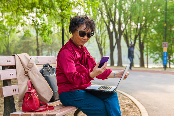 Vieja mujer asiática usando teléfono móvil y oliendo en el jardín en el parque. Feliz vieja mujer de negocios asiática usando el teléfono inteligente y el ordenador portátil en el parque de verano
. - Foto, imagen