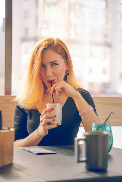pretty happy redheaded caucasian woman in black shirt drinks cup of coffee in the modern cafe alone - Фото, зображення
