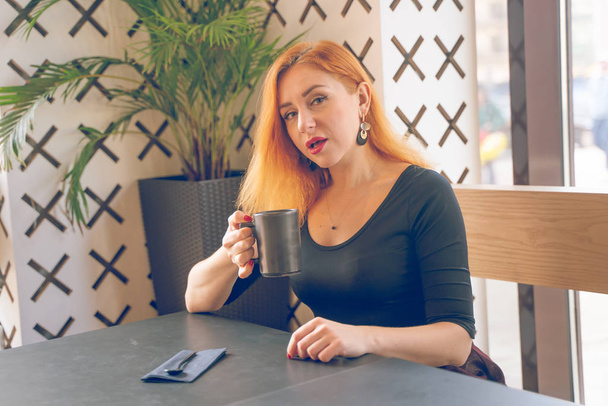 mujer caucásica pelirroja bastante feliz en camisa negra bebe taza de café solo en la cafetería moderna
 - Foto, imagen
