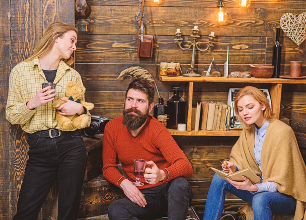 Woman absorbed by interesting book and not noticing her family. Girl with teddy bear in retro outfit standing next to wooden wall. Bearded man with cheerful look enjoying time with wife and daughter - Foto, imagen