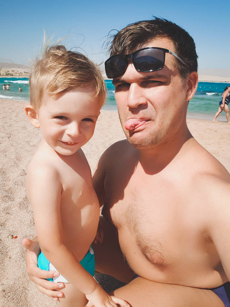 Zabawny wizerunek młodego mężczyzny z jego małego syna pokazując językami w aparacie podczas robienia zdjęć selfie na plaży morskiej. Rodzinny relaks i dobry czas podczas letnich wakacji. - Zdjęcie, obraz